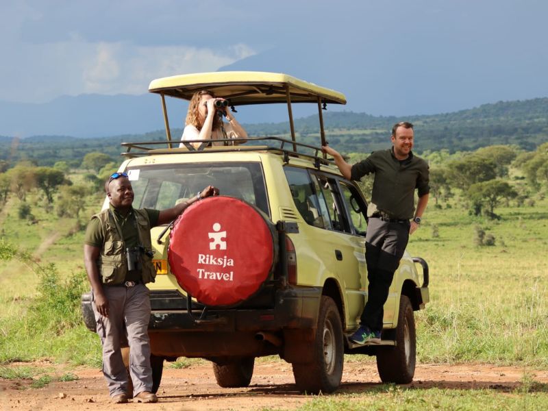 Safarivoetuig met open dak staat in een groene savanne, een vrouw kijkt door een verrekijker terwijl twee mannen naast de auto staan