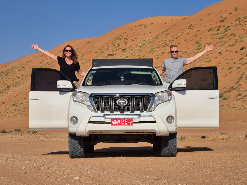 Een man en vrouw staan in de open portieren van een witte Toyota Land Cruiser, geparkeerd in een woestijnlandschap met  hoge oranje zandduinen op de achtergrond. Beiden hebben hun armen wijd uitgespreid en lachen