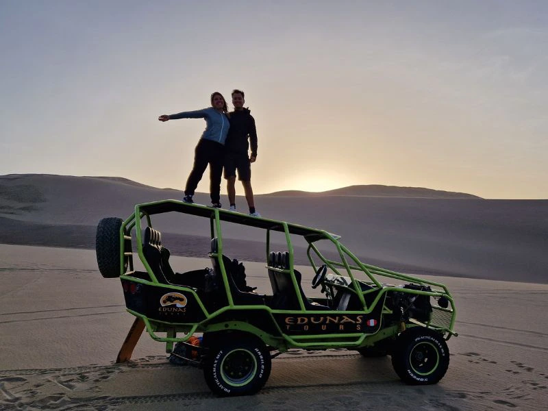 Een man en vrouw staan bovenop een felgroene buggy in een uitgestrekt duinlandschap bij zonsondergang, met de woestijnheuvels op de achtergrond.