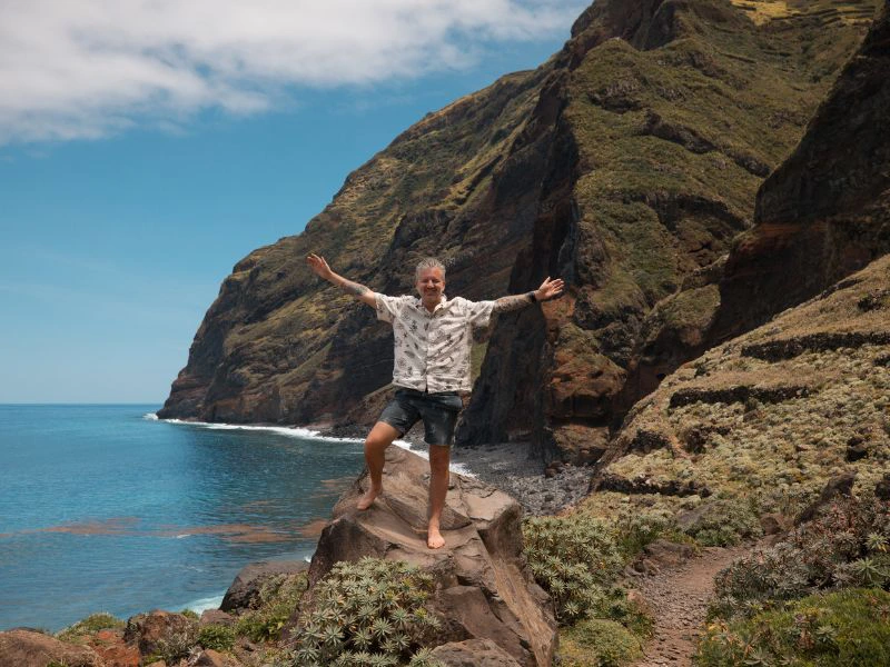 Man staat met open armen op een rots langs de kust, met steile groene kliffen en de oceaan op de achtergrond.