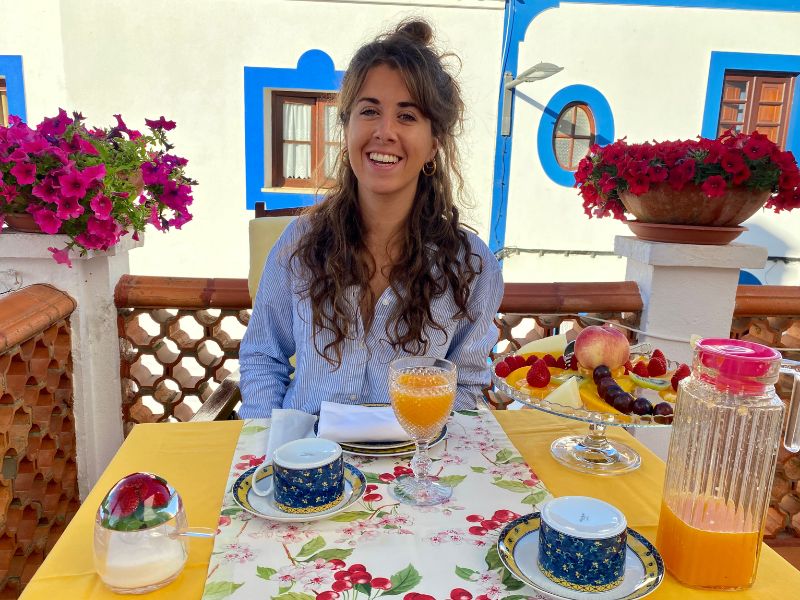 Vrouw zit lachend aan een kleurrijk gedekte ontbijttafel met bloemen, sap en fruit op een zonnig terras met witte muren en blauwe kozijnen