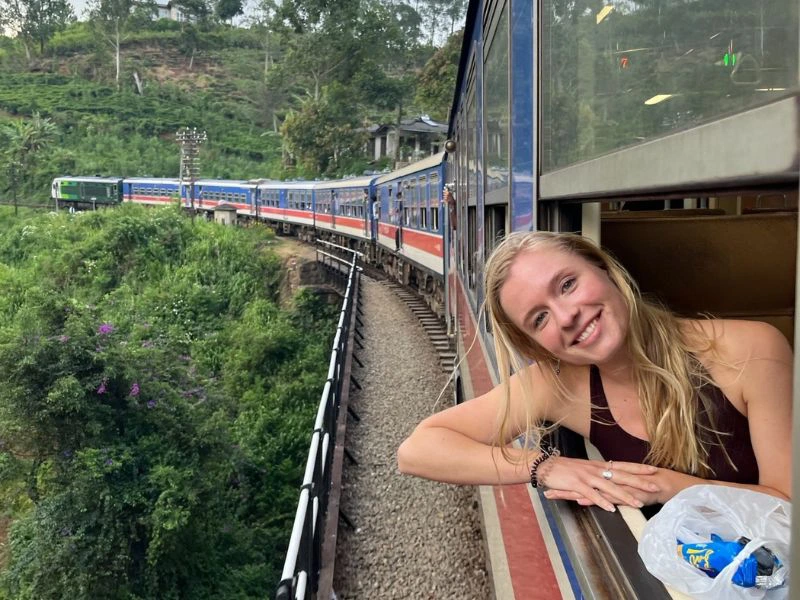 Vrouw leunt lachend uit het raam van een kleurrijke trein die door een groene, heuvelachtige omgeving rijdt.