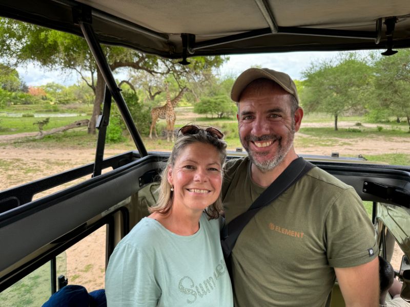 Man en vrouw glimlachen in een safarivoertuig, met op de achtergrond een giraffe in de groene safari