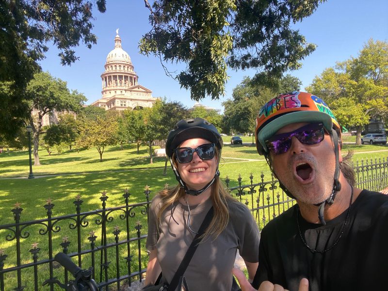 Twee mensen maken een vrolijke selfie met fietshelmen op, met het Texas State Capitol op de achtergrond op een zonnige dag