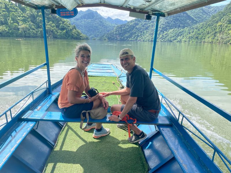 Een man en vrouw zitten glimlachend naast elkaar op een blauw bankje in een open boot, varend over een rustig groen meer, omringd door groene bergen en jungle
