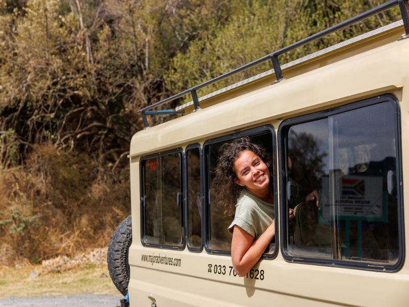 Vrouw leunt lachend uit het raam van een beige safaribus in een droge, berachtige omgeving