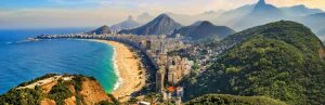 Panoramisch uitzicht op de kust van Rio de Janeiro met de lange boog van Copacabana Beach, de oceaan aan de linkerkant en groene heuvels en stadsbebouwing tegen de achtergrond van grillige bergen.