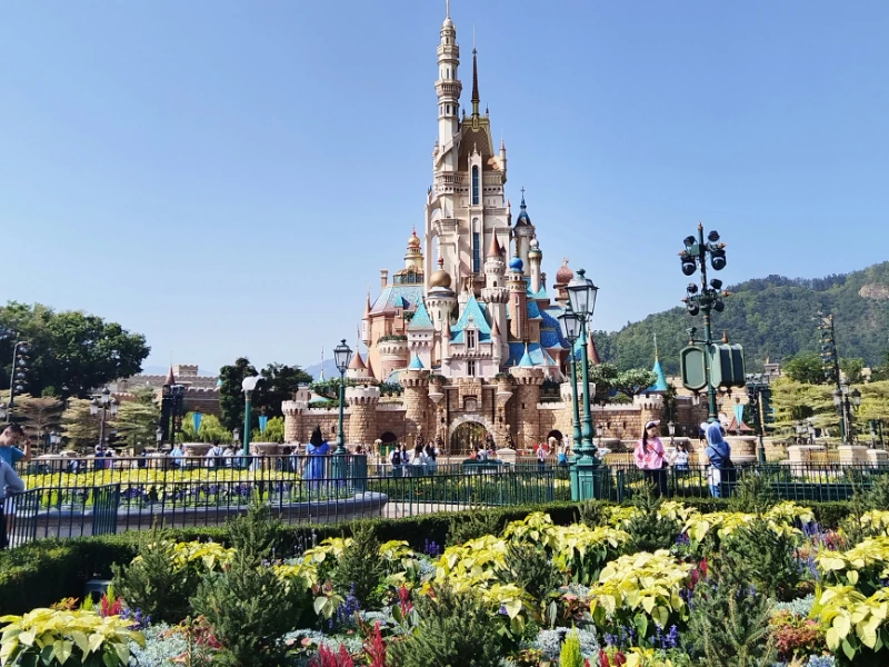 Disney kasteel in Disnelyland Hong Kong tegen een strak blauwe lucht en groene planten en kleurrijke bloemen op de voorgrond.