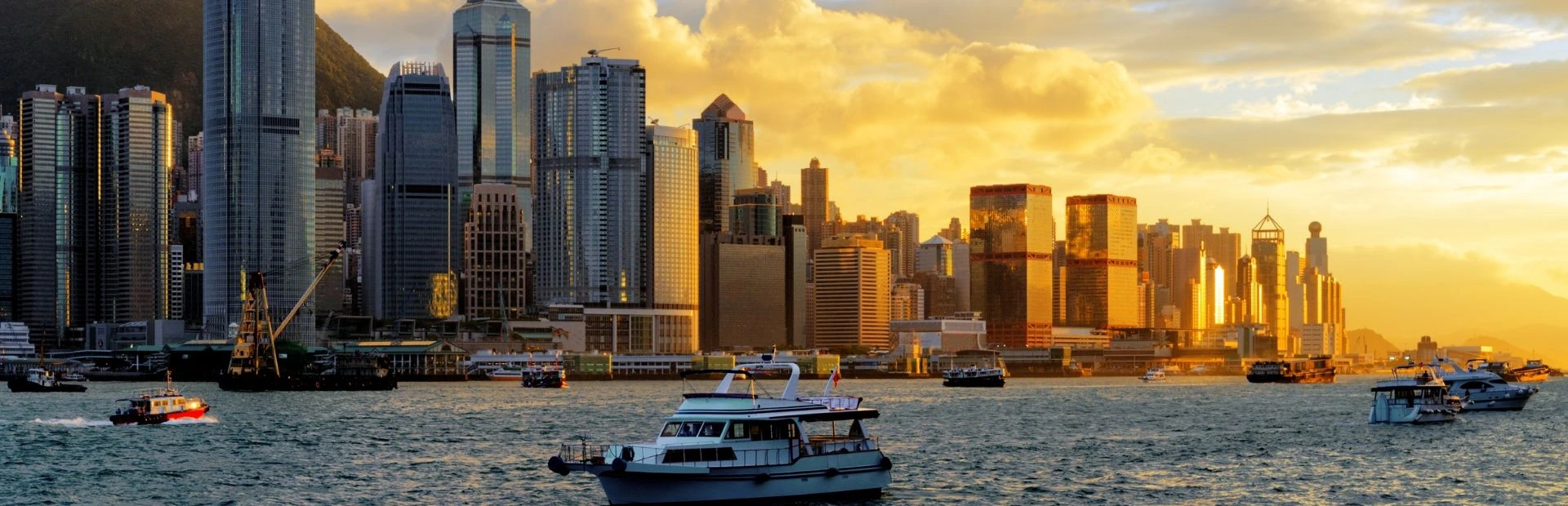 Je ziet de skyline van Hong Kong met bootjes en het water op de voorgrond. De foto is genomen tijdens de zonsondergang waardoor de oranje kleuren reflecteren op de ramen van de hoge gebouwen.
