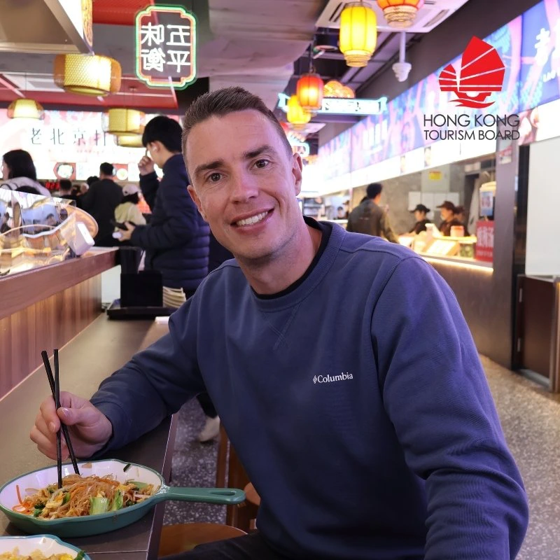 Man met blauwe trui aan in een restaurant in Hong Kong die een kom noedels zit te eten.