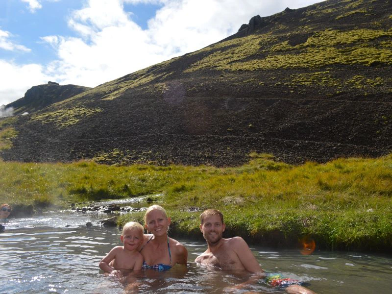 Gezin met kind zit lachend in een natuurlijke warmwaterbron in een berglandschap.