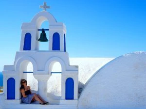 Vrouw zit onder de witte en blauwe klokkentoren van een Grieks kerkje op Santorini, met een helderblauwe lucht op de achtergrond.