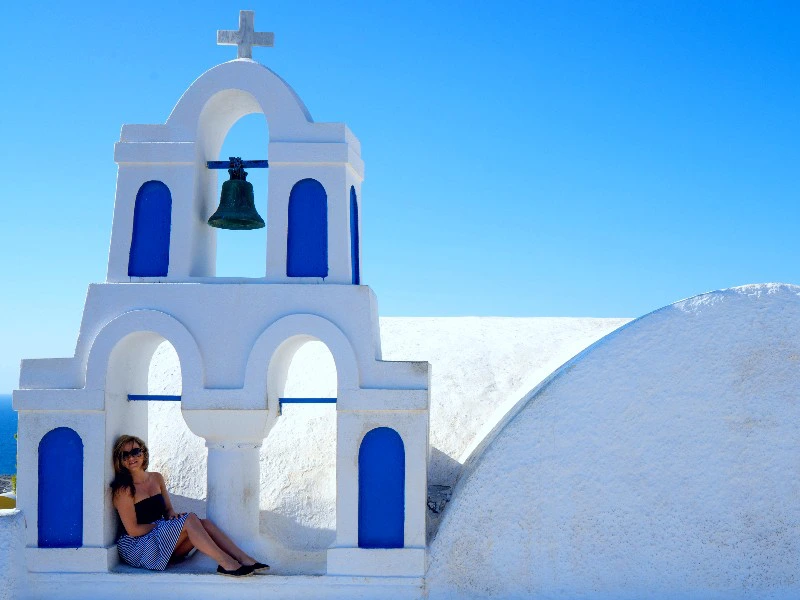 Vrouw zit onder de witte en blauwe klokkentoren van een Grieks kerkje op Santorini, met een helderblauwe lucht op de achtergrond.
