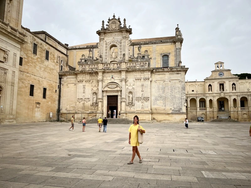 Vrouw in gele jurk staat op een plein voor een barokke kathedraal met rijk versierde gevel.