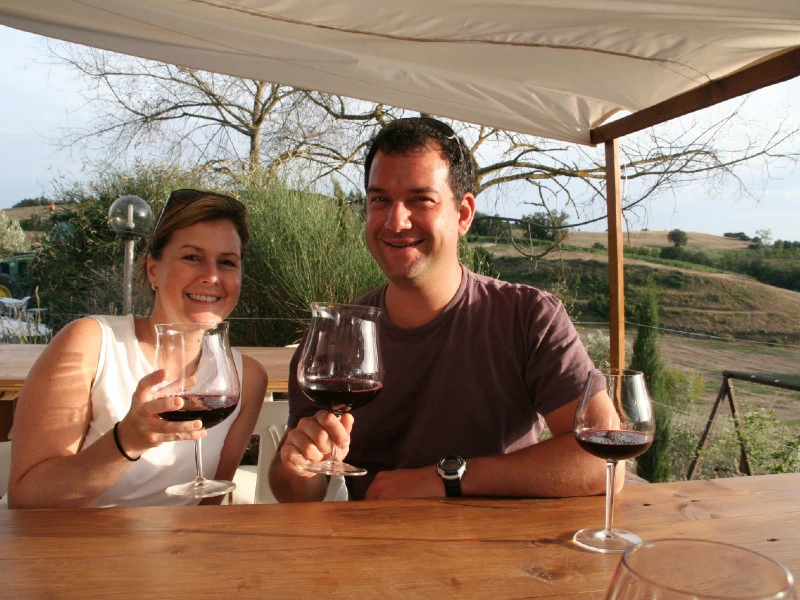 Man en vrouw zitten buiten aan een houten tafel en proosten met glazen rode wijn.