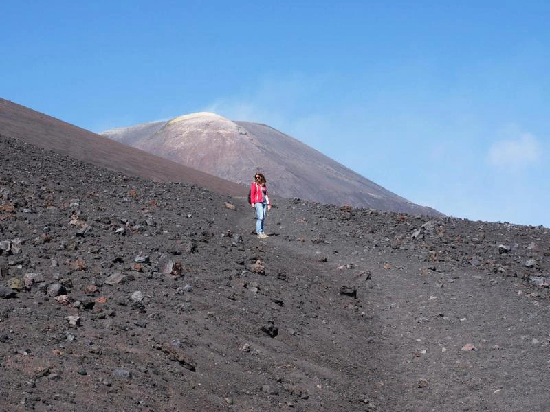 Vrouw wandelt over een zwart lavalandschap met de rokende top van een vulkaan op de achtergrond.