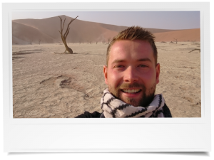 Selfie van een man in de woestijn van Deadvlei in Namibië, met achter hem de witte kleivlakte, dorre bomen en hoge oranje zandduinen.