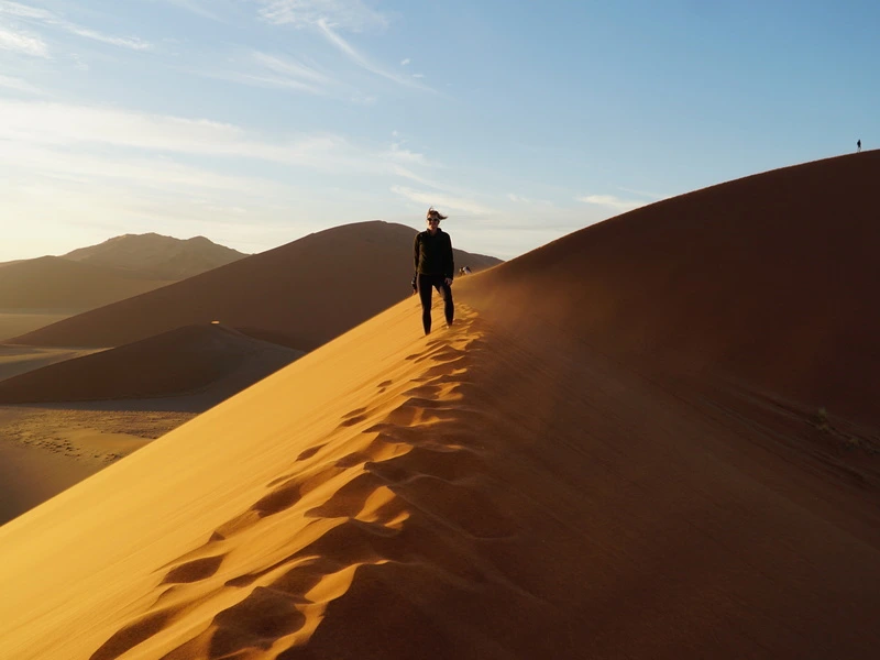 Een persoon loopt over de top van een hoge zandduin in de woestijn van Namibië, met een rij voetstappen achter zich en de zon die zacht over het zand schijnt.