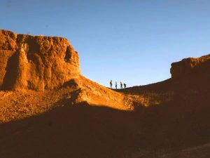 Vier silhouetten van wandelaars staan bovenop een rotsachtige bergkam in een woestijnlandschap, verlicht door de warme gloed van de avondzon.