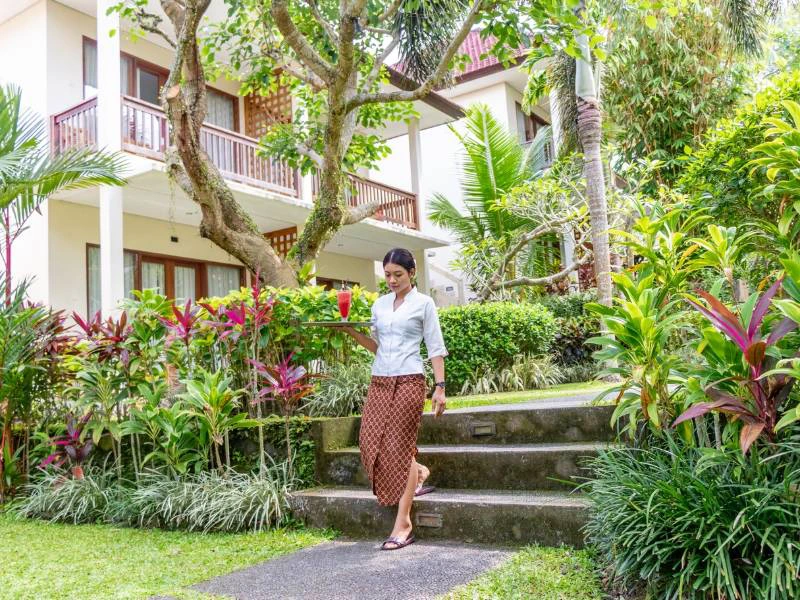 Vrouw in traditionele kleding die door een tropische tuin loopt bij een kleinschalig hotel met balkons en veel groen.