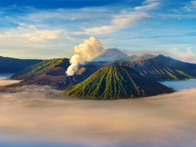 Luchtfoto van de Bromo-vulkaan op Java, Indonesië. In het midden van het beeld zie je de actieve krater van de Bromo, waar witte rook uit opstijgt. De vulkaan zelf heeft steile, groene ribbelvormige hellingen. Rondom ligt een uitgestrekte zandvlakte en een dikke laag ochtendmist of wolken die laag over het landschap hangt. Op de achtergrond rijzen andere vulkanische pieken op, waaronder de Semeru in de verte. De lucht is helderblauw met enkele hoge wolken.