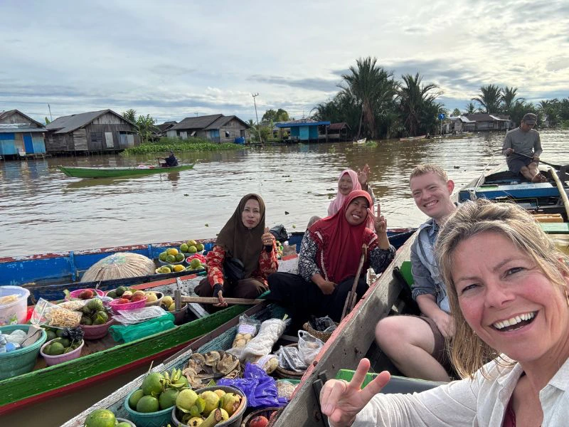 Selfie van een vrouw op een drijvende markt in Kalimantan, omringd door lokale verkopers in kleurrijke boten vol groenten, fruit en andere waren.