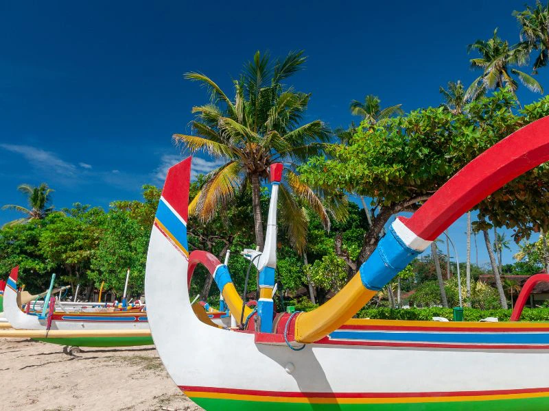 Kleurrijke traditionele vissersboot op een tropisch strand in Sanur Bali, omringd door palmbomen en helderblauwe lucht.