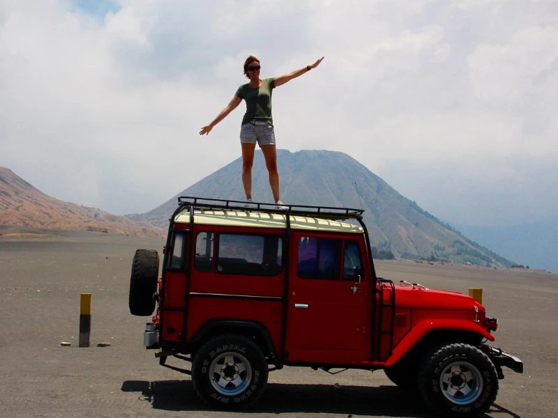 Een vrouw staat op een rode Jeep met haar armen wijd. Links van de auto staat een klein paaltje en achter de vrouw zie je de Bromo vulkaan en een lichtblauwe lucht met witte wolken.