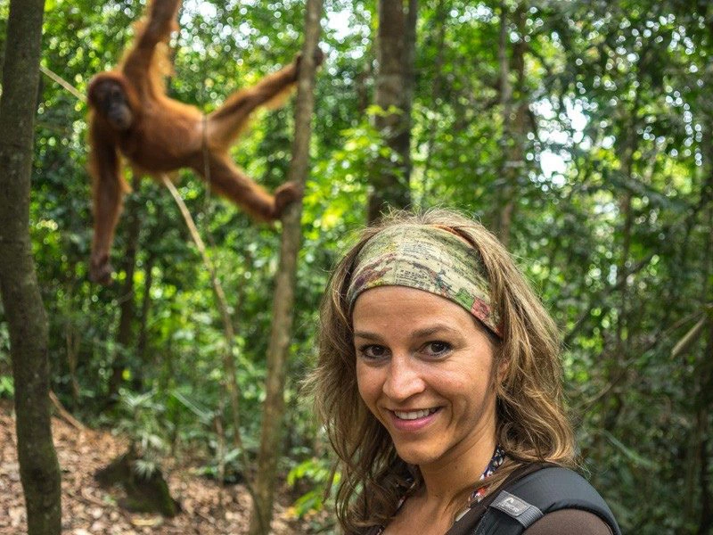 Een reizigster met bruin haar en een haarband om haar hoofd maakt een selfie met een wazige orang oetan links achter haar. Ze staat in een dichtbebost stukje jungle in Sumatra.