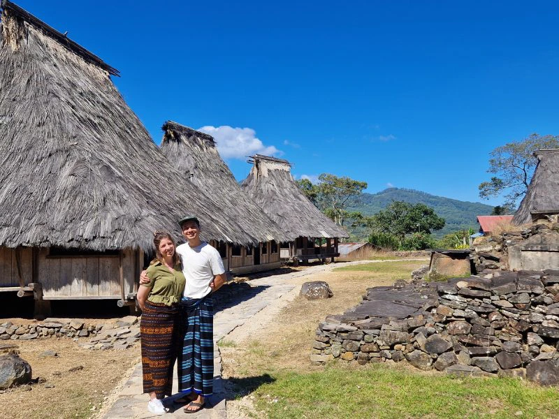 Een stel staat naast elkaar met sarongs om hun middel voor houten en rieten hutjes van een traditioneel dorp in Flores en naast hen stapels met stenen in het gras. In de verte zie je het bergachtige landschap en de blauwe lucht met een enkele wolk.