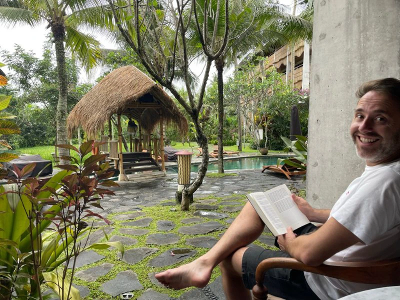 Een man zit ontspannen in een houten stoel, leest een boek en kijkt uit over een tropische tuin met een zwembad, palmbomen en een rieten gazebo. Het geheel straalt rust en vakantiegevoel uit; vrolijke groene planten sieren het terras met natuurstenen tegels.
