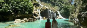 Twee vrouwen vanaf hun achterkant dit kijken naar het helderblauwe water met rotsen en een grote waterval. De blauwe lucht komt een klein beetje door de groene bomen en struiken op de rotsen door.