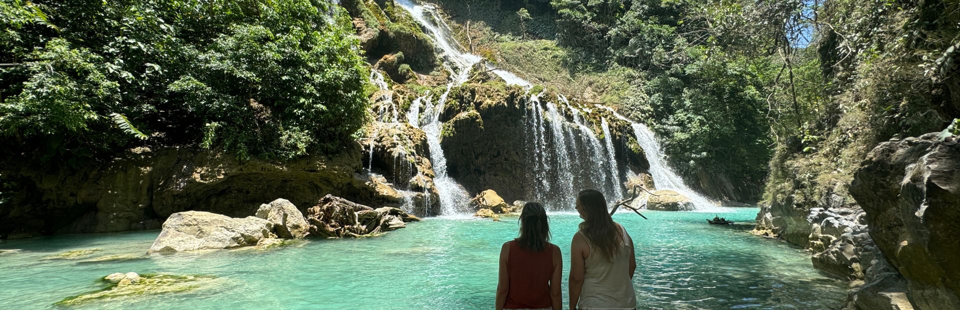 Twee vrouwen vanaf hun achterkant dit kijken naar het helderblauwe water met rotsen en een grote waterval. De blauwe lucht komt een klein beetje door de groene bomen en struiken op de rotsen door.