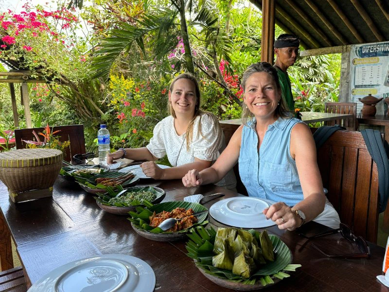 Twee mensen zitten aan een houten tafel in een tropische tuin en genieten van een traditionele Indonesische maaltijd met verschillende gerechten geserveerd op bananenblad. Op de achtergrond veel groene planten en felgekleurde bloemen, terwijl de sfeer ontspannen en gezellig is.