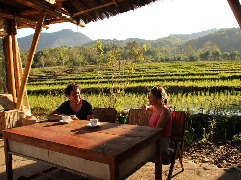 Twee reizigers drinken koffie aan een houten tafel onder een rieten dak, met uitzicht op zonovergoten rijstvelden op Flores, Indonesië. Op de achtergrond liggen groene heuvels in het zachte ochtendlicht.