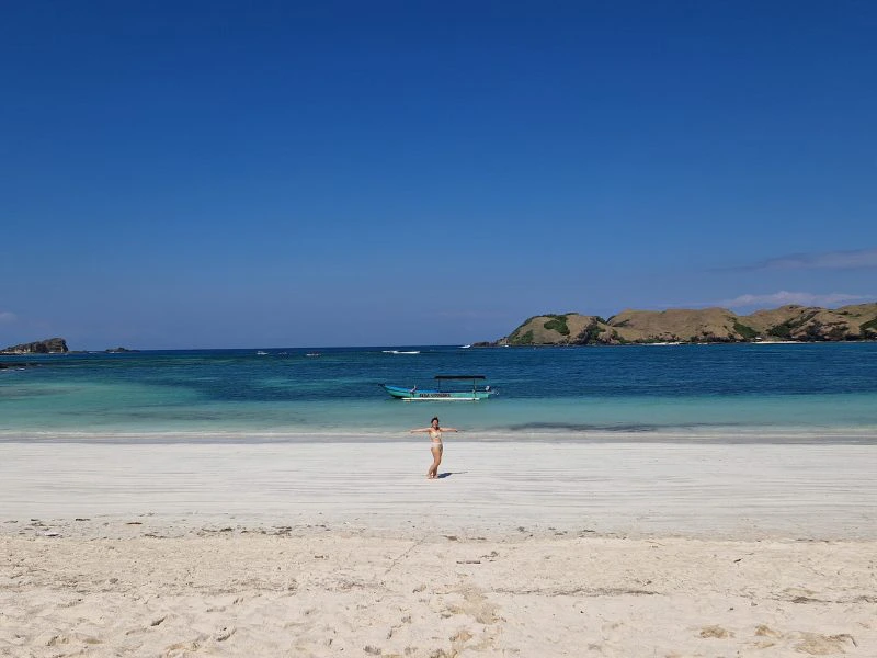 Een reizigster in bikini staat dichtbij de zee met haar armen wijd. Het strand is van licht zand en de zee kleurt van turquoise tot aan donkerblauw en daarachter zijn heuvels een een blauwe lucht te zien.