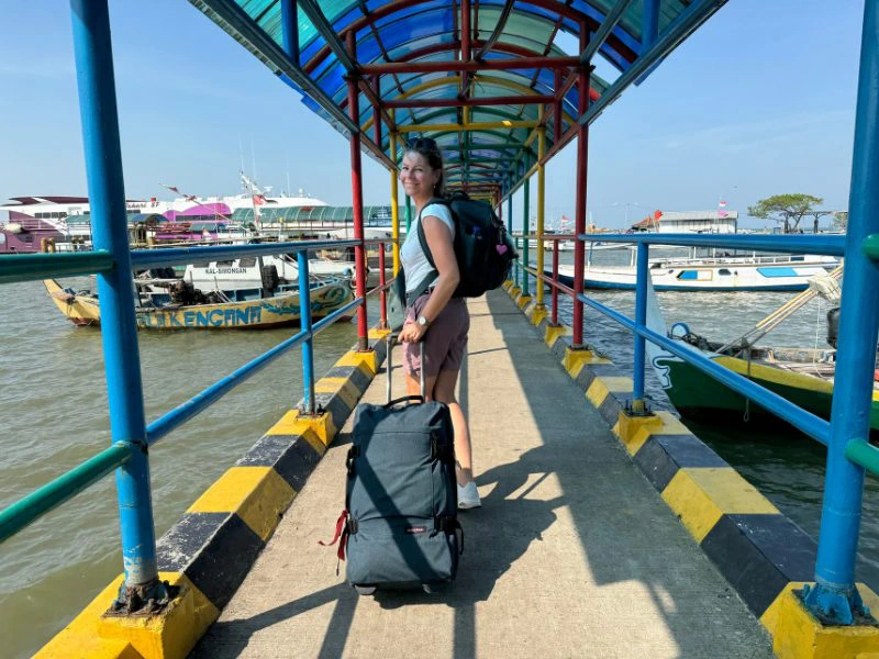 Reizigster met rugzak en reistas loopt over een kleurrijke, overdekte pier in Karimunjawa, Indonesië. Ze kijkt lachend achterom. Links en rechts liggen boten in het water, de zon schijnt fel en de lucht is helderblauw.