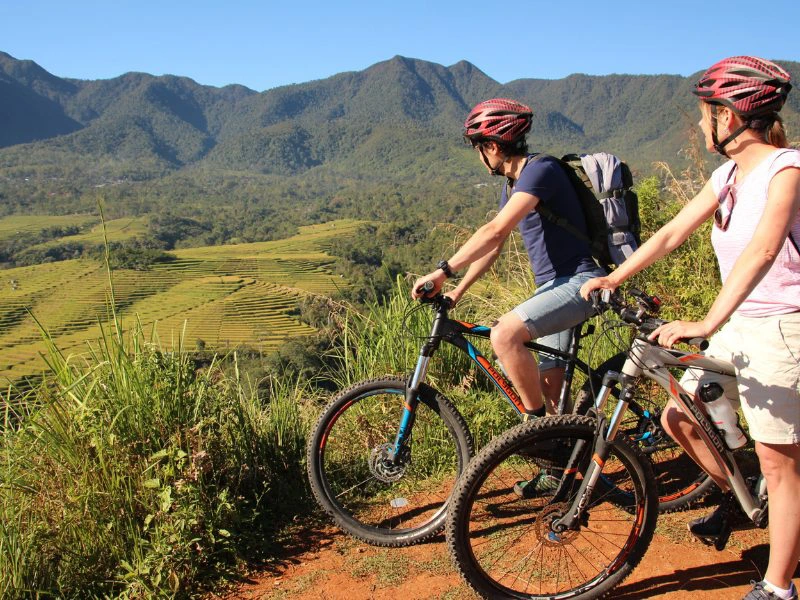 Twee reizigers op mountainbikes met rode helm op kijken naar het uitzicht op de rijstterrassen in Ruteng met groene heuvels in de verte.