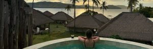 Een vrouw ontspant in een ronde buitenjacuzzi en kijkt uit over een tropisch eilandlandschap met rieten bungalows, palmbomen en een rustig zeezicht bij zonsondergang. Op de rand van de jacuzzi staat een glas donkere drank.
