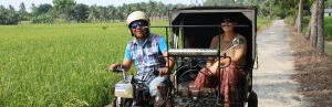 Een lachende TukTuk driver met witte helm op rijdt tussen het groen en de palmbomen in de verte met een reizigster erin in Medan op het eiland Sumatra.