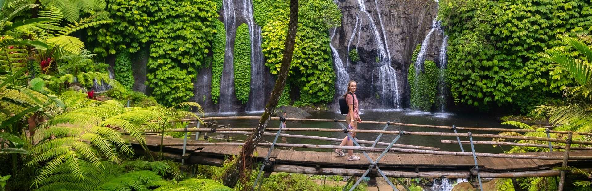 Reiziger op Bali bij een waterval rondom het plaatsje Munduk. Ze loopt over een bamboe-brug met op de achtergrond een waterval. Het is omringd door groene planten en bomen.