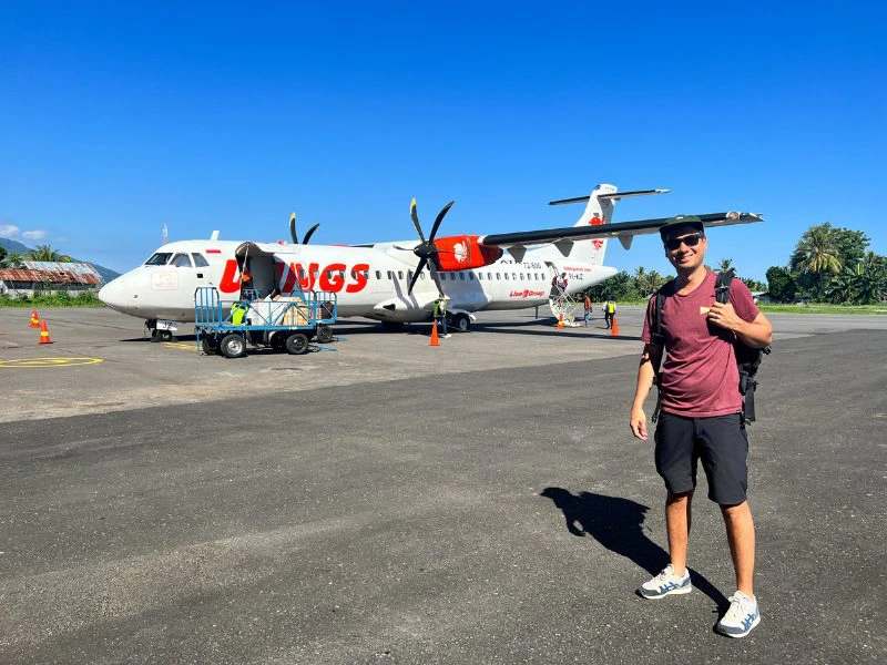 Man met rugzak en pet staat op het vliegveld voor een wit-oranje propellervliegtuig van Wings Air. Het is een zonnige dag met een blauwe lucht. Op de achtergrond zie je tropische bomen en een klein gebouw. De man draagt een bordeauxrode t-shirt, zwarte shorts en witte sneakers.