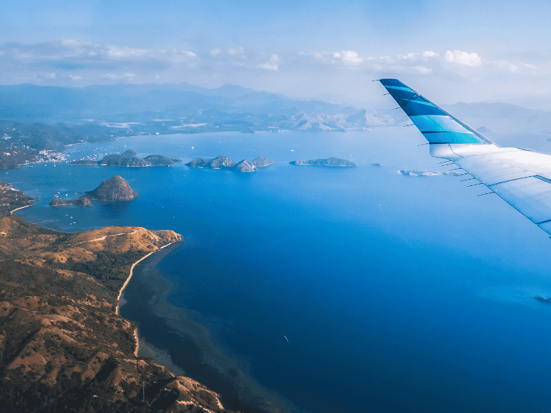 Luchtfoto van een vliegtuig boven de kustlijn van een Indonesisch eiland. Rechts zie je de vleugel met blauwe en witte strepen. Onder het toestel ligt een uitgestrekte, diepblauwe zee met hier en daar kleine witte bootjes. Links is een ruige, heuvelachtige kust zichtbaar die overgaat in een baai met kleinere eilanden met steile kliffen. Aan de horizon doemt een bergachtig landschap op, gehuld in een lichte waas. De lucht is licht bewolkt met hoge wolken.