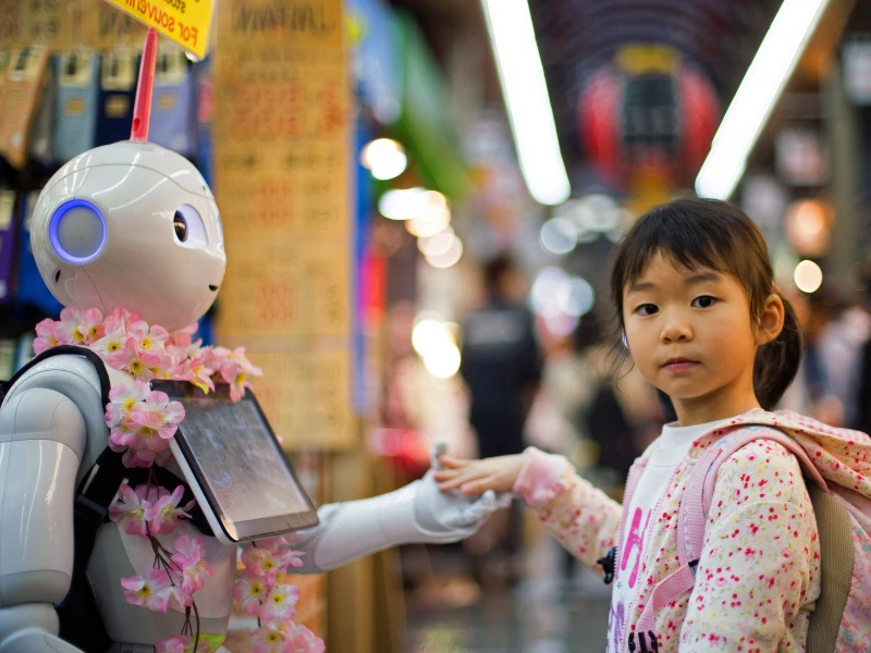 Een jong meisje kijkt in de camera terwijl ze de hand vasthoudt van een humanoïde robot met een bloemenkrans en scherm op de borst, in een levendige marktstraat in Japan.