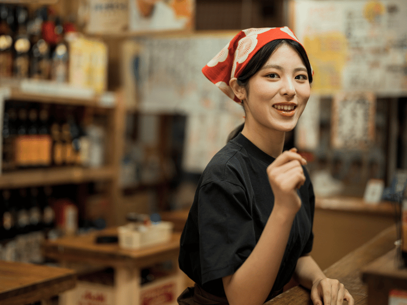Jonge Japanse vrouw met rood-witte hoofddoek glimlacht achter een bar in een Japanse eetgelegenheid.