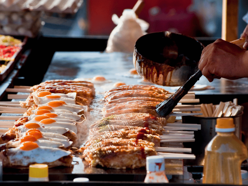 Close-up genomen tijdens een foodtour van een grill in Fukuoka, Japan, waar een kok spiesjes met eieren en andere ingrediënten bestrijkt met saus.