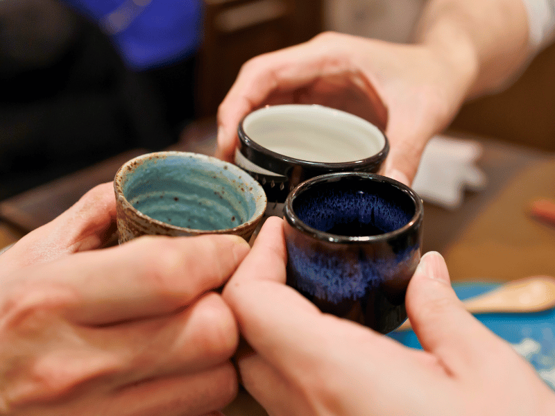 Drie handen die kleine keramische kopjes bij elkaar houden met sake voor een toast tijdens de fukuoka food tour