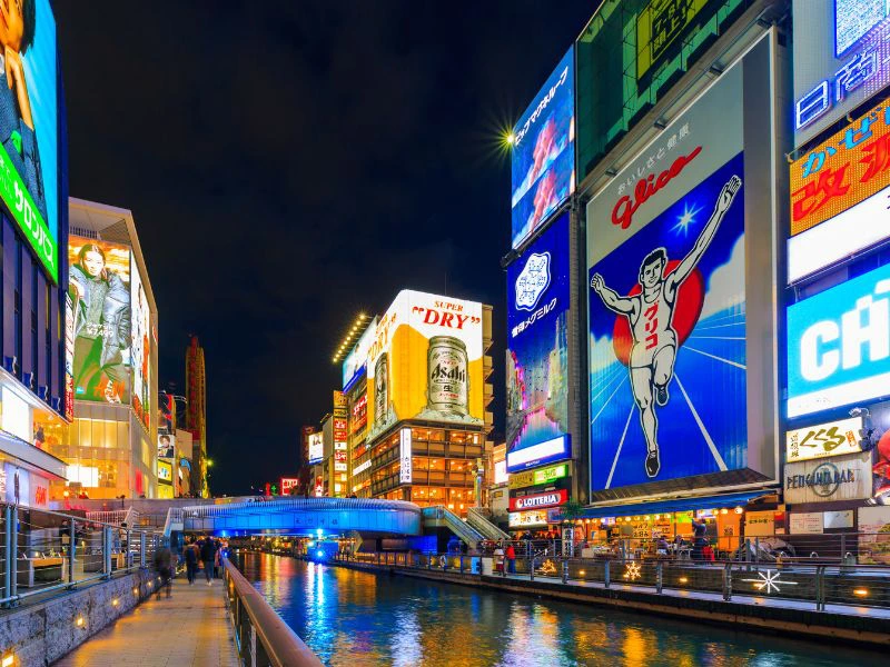 Avondbeeld van de Dotonbori-wijk in Osaka met kleurrijke neonreclames en billboards langs het kanaal, waaronder het bekende Glico-man bord.