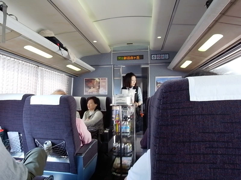 Binnenkant van een Japanse hogesnelheidstrein, waar een stewardess met een servicekar drankjes en snacks langs de zitplaatsen aanbiedt aan passagiers.