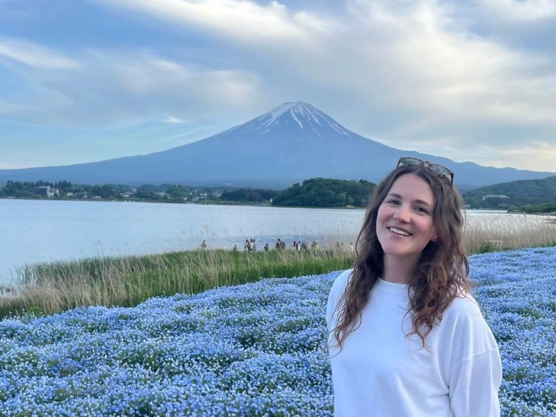 Vrouw staat lachend in een veld met blauwe bloemen aan de oever van een meer, met Mount Fuji op de achtergrond.